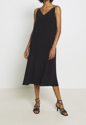 Kvinde iført en sort midi-kjole uden ærmer med dobbelte tynde stropper og sorte sandaler med hæl og stropper, stående foran en ensfarvet baggrund.