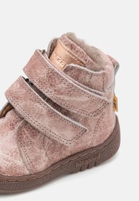 Bottine en daim rose avec deux sangles Velcro, semelle en caoutchouc marron, et accents dorés. Finition texturée avec un intérieur doux et moelleux.