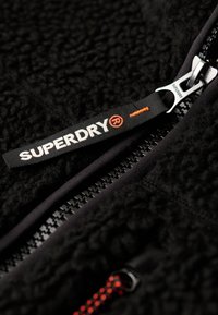 Zwarte fleece jacket met een gestructureerde oppervlakte, uitgerust met een zilveren rits en een label met de tekst "SUPERDRY" in wit met een oranje accent.
