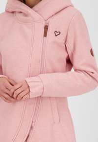 Giacca in pile rosa con chiusura a zip, caratterizzata da un logo a forma di cuore e un accento marrone sulla manica. Texture morbida e design con cappuccio.