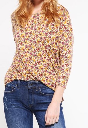 Femme portant un haut à manches longues à motifs floraux dans des tons jaunes et rouges, associé à un jean bleu, debout avec une main détendue le long du corps.