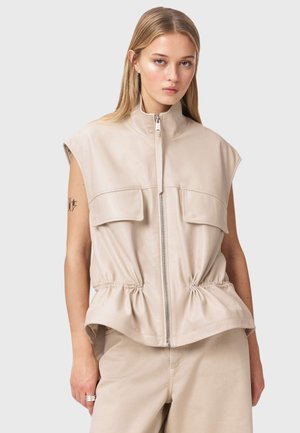 Nuori nainen, jolla on pitkät vaaleat hiukset, pukeutuneena hihattomaan beigeen vetoketjuliiviin, jossa on etutaskut, ja beigen värisiin housuihin yksiväristä taustaa vasten.