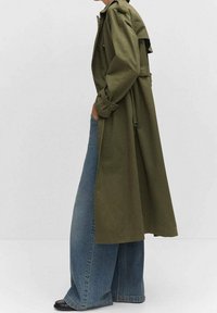 Długi oliwkowozielony płaszcz z paskiem, z wysokim kołnierzem i bocznymi kieszeniami, połączony z szerokimi niebieskimi dżinsowymi spodniami i czarnym obuwiem.
