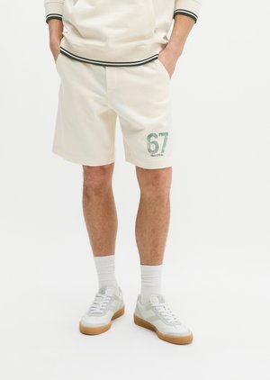 Mężczyzna w kremowych szortach z nadrukiem "67" na lewej nodze, białe skarpetki, białe trampki i kremowa bluza z kapturem z czarną lamówką, stoi z rękami w kieszeniach.
