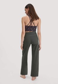 Vrouw die blootsvoets staat, met haar rug naar ons toe, gekleed in een donkere wijde broek en een zwart-roze gestreepte cropped top met kruislings straps.