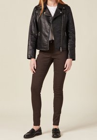 Chaqueta de moto negra de piel sintética con cierre de cremallera y herrajes plateados, combinada con pantalones de tela acanalada marrones y mocasines negros.