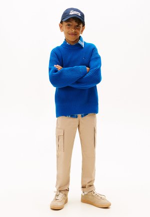 Maglione blu lavorato a maglia, camicia a scacchi sotto, pantaloni cargo beige con tasche e scarpe da ginnastica chiare. Cappellino nero con logo "Tommy".