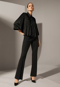 Vrouw in een losse zwarte geplooide blouse met pofmouwen, zwarte slim-fit broek met witte stippen en zwarte puntige hakken, die zelfverzekerd staat.