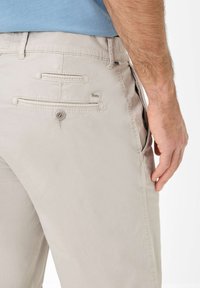 Lichtbeige broek met een gladde textuur, voorzien van achterzakken met kleppen en een klein logo label. Een subtiele knoopsluiting is zichtbaar.