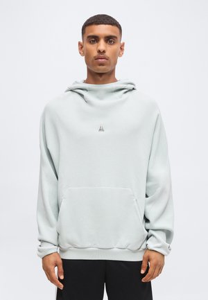Jeune homme portant un sweat à capuche gris clair texturé avec un petit logo sur la poitrine et un pantalon noir, debout devant un fond uni.