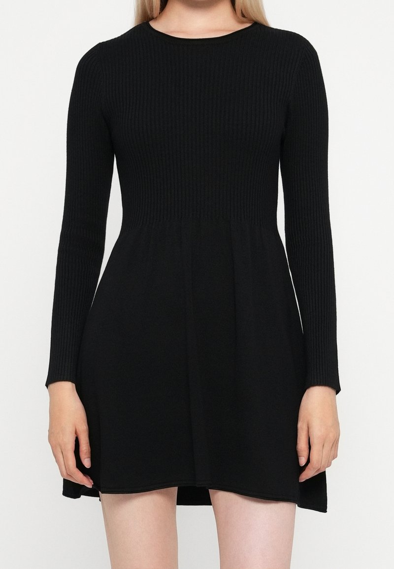 Femme portant une robe noire à manches longues côtelée avec une jupe évasée, debout devant un fond blanc uni.