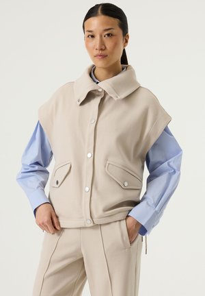 Woman standing with hands in pockets, wearing a beige vest and pants over a light blue shirt, neutral expression, light background.