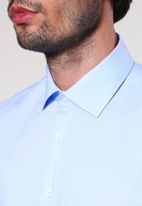 Ljusblå skjorta med knäppning och klassisk krage, med en mjuk textur och subtil lyster. Inkluderar vita knappar på framsidan.