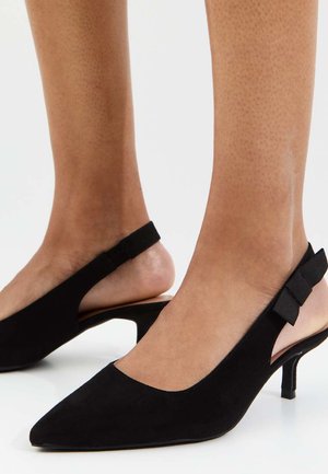 Zwarte suède pumps met spitse neus en lage kittenhiel, gedragen op blote voeten tegen een effen achtergrond.