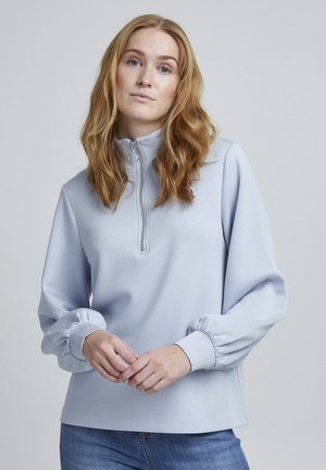 Lys blå zip-up sweatshirt med høj krave, der har plissede ærmer og en blød tekstur, sat sammen med blå jeans.