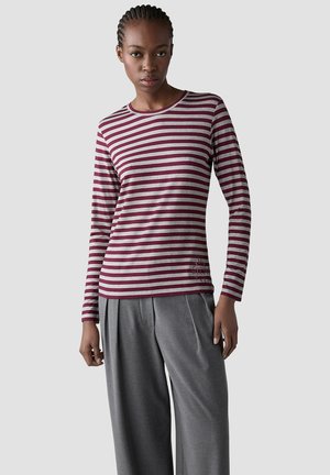 Woman wearing a long-sleeve burgundy and gray striped shirt paired with loose gray pants, standing against a plain light gray background.
