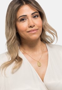 Gold pendant necklace with oval charm, engraved detail, fine chain, worn over a light-colored, smooth fabric top. Subtle shine and texture.