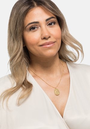 Goldene Anhänger-Halskette mit ovalem Charm, gravierten Details, feiner Kette, getragen über einem hellen, glatten Stoffoberteil. Subtile Glanz und Struktur.