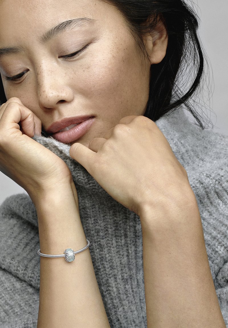 Strieborný náramok s textúrovaným okrúhlym zapínaním, nosený na holom zápästí. Modelka má na sebe mäkký šedý pletený sveter.
