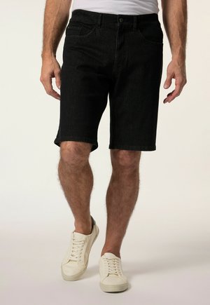 Mann trägt schwarze Denim-Shorts, weiße Sneakers und ein weißes T-Shirt, steht vor einem einfarbigen hellen Hintergrund.