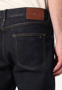 Tmavomodré džínsy z denimu majú hnedú koženú nášivku s vyrazeným nápisom "Tommy Hilfiger", kontrastné žlté prešívanie a zadné vrecká so subtílnym brandingom.
