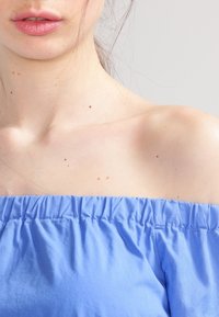 Top azzurro chiaro scollato sulle spalle con scollatura arricciata, texture liscia e tessuto morbido, che mette in mostra le spalle scoperte e una parte del collo.