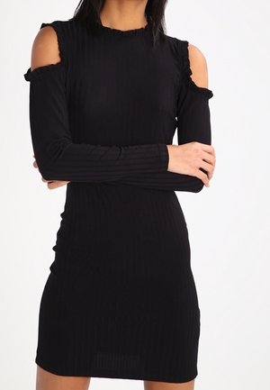 Frau trägt ein eng anliegendes schwarzes geripptes Kleid mit Cut-Outs an den Schultern und langen Ärmeln, steht mit verschränkten Armen.