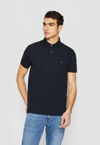 Dunkelblaues Polohemd mit klassischem Kragen, kurzen Ärmeln und kleinem Logo auf der Brust. Der Stoff hat eine glatte Textur. Getragen mit hellblauen Jeans.