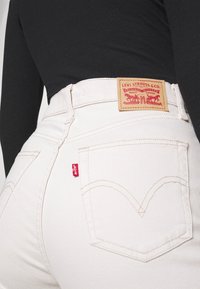 Jeans de denim blanco con una cintura alta, que presentan una etiqueta de cuero, una etiqueta roja y costuras clásicas en los bolsillos traseros. Diseño de pierna recta.