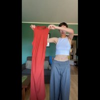 Vestido vermelho sem mangas com uma textura canelada, sustentado, ao lado de uma pessoa com uma blusa branca curta e calças largas cinzas. Fundo de parede verde.
