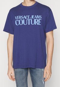 Camiseta de algodón azul marino con mangas cortas, que presenta "VERSACE JEANS COUTURE" en texto grueso de color azul claro en el pecho. Textura suave.