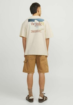 Beige katoenen t-shirt met grafische print op de rug, gecombineerd met bruine cargoshorts. Het model heeft donker haar en draagt beige en bruine schoenen.