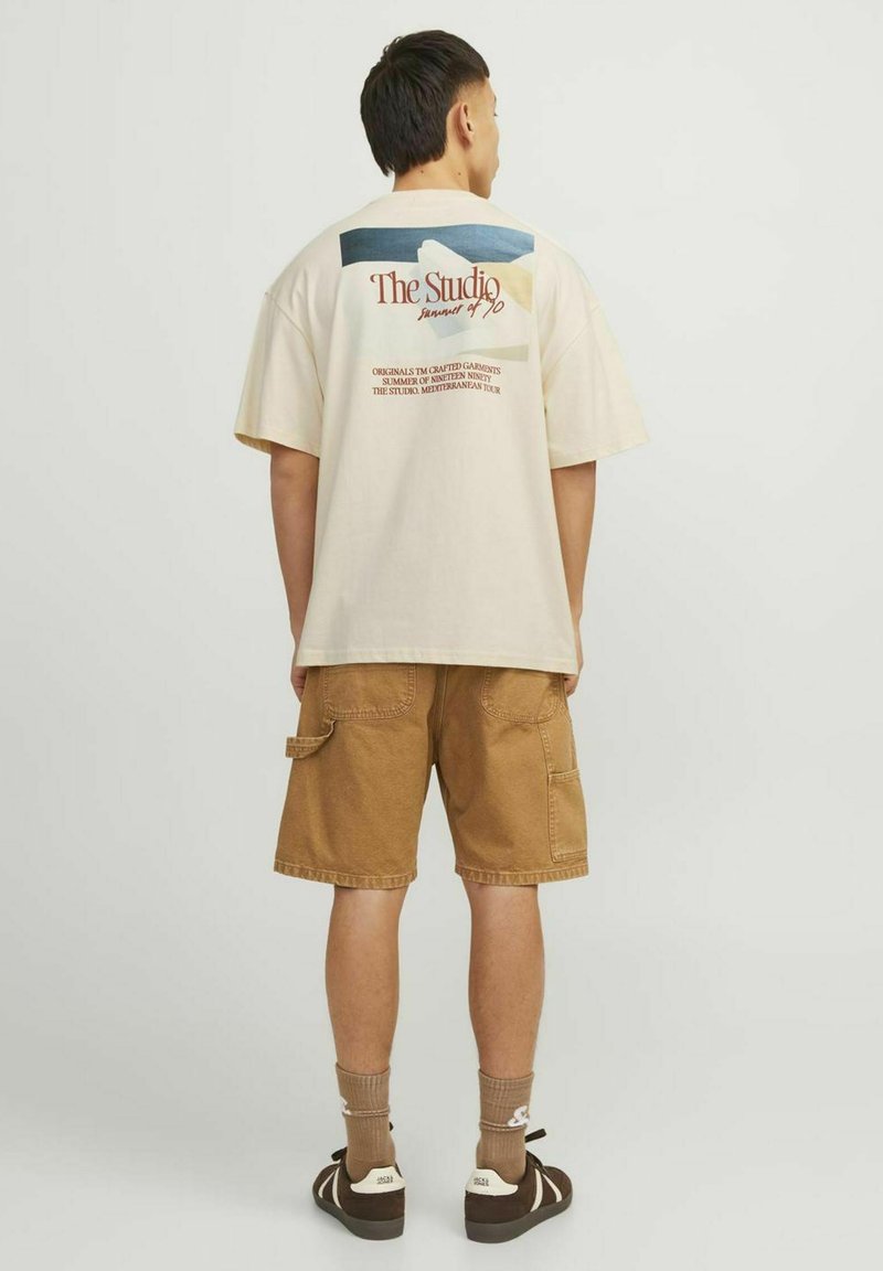 Beige katoenen t-shirt met grafische print op de rug, gecombineerd met bruine cargoshorts. Het model heeft donker haar en draagt beige en bruine schoenen.
