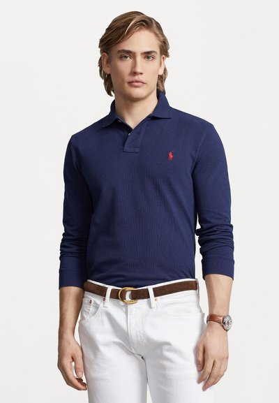 Marineblaues Langarm-Polohemd mit rotem Logo auf der Brust, kombiniert mit weißen Hosen und einem braunen Ledergürtel. Strukturierter Stoff.