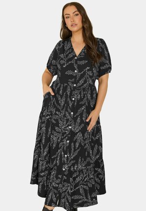 Frau trägt ein schwarzes, knöpfbares Midikleid mit weißen Blattmustern, kurzen Ärmeln und Taschen, steht mit einer Hand in der Tasche und der anderen auf dem Rock.