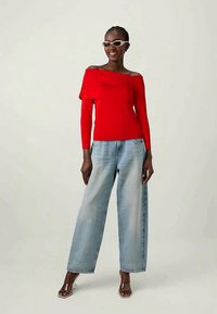 Top vermelho sem ombros com detalhe de tecido franzido, combinado com jeans de perna larga em lavagem clara e sandálias de salto transparente.