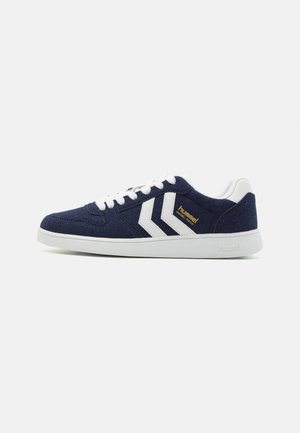 Baskets bleu marine avec lacets blancs, semelle blanche, bandes chevrons blanches et logo "hummel" doré sur le côté.