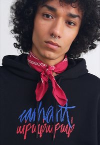 Felpa nera con un design di testo multicolore in rosso e blu. Un fazzoletto rosso con motivi bianchi legato attorno al collo. Capelli ricci in mostra.