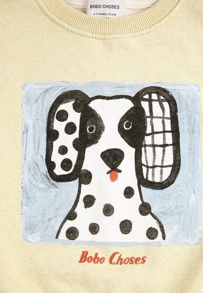 Kreslený dalmatín pes s jedným uškom škvrnitým a druhým kockovaným, vyplazeným jazykom, na svetložltom látkovom pozadí s textom „Bobo Choses“.