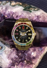 Goldene Timex-Uhr mit Regenbogen-Edelsteinen rund um das schwarze Zifferblatt mit einem Augendesign, liegend auf violetten Amethystkristallen.