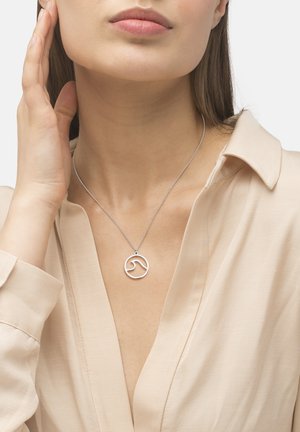 Zilveren hanger ketting met golvenontwerp, ronde vorm, hangend aan een fijne ketting, gedragen over een lichtbeige blouse met een diepe halslijn.
