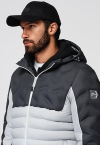 Svart och grå uppblåsbar jacka med dragkedja fram, texturerade paneler och huva, lager på lager över en svart hoodie. Har en logotyp på ärmen.