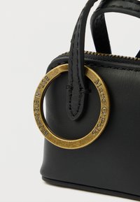 Canto de uma mala de couro preta com uma etiqueta metálica circular dourada gravada com "Marc O’Polo" e "EST. IN STOCKHOLM", alças pretas costuradas e fecho de correr visível.