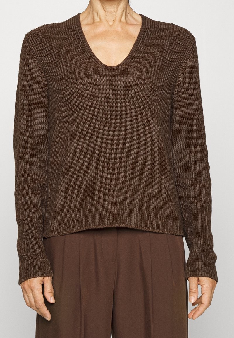 Personne portant un pull marron en maille à manches longues avec col en V et un pantalon marron plissé assorti, mains détendues le long du corps.