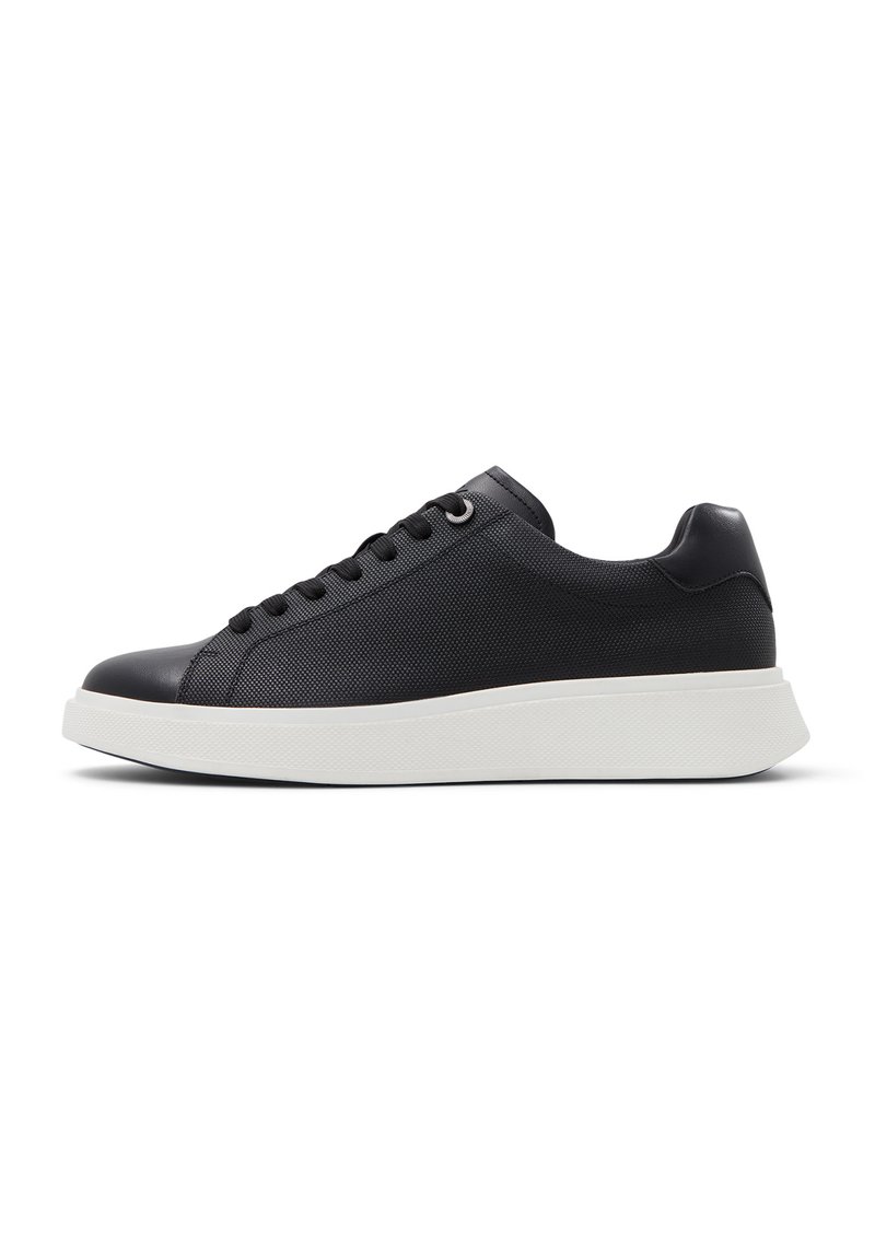 Svart sneakers med teksturert overdel, rund tå og flat hvit såle. Har runde snørebånd og dekorative snøløkker. Minimalistisk design.