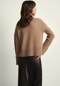 Brązowy sweter wykonany z wolno-blendowanej wełny, krótkiej długości, z prążkowanym wykończeniem u dołu i na mankietach, ukazujący luźny krój i miękką strukturę. Nosi się go na ciemnej, zwiewnej spódnicy.