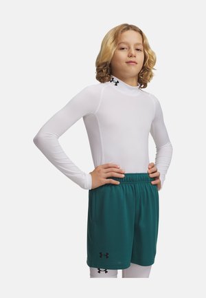 Blond dítě stojí s rukama v bok oblečené do bíle dlouhého kompresního trička, tyrkysových sportovních šortek a bílých legín s logy.