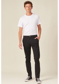 Schwarze Baumwollchinos mit schlanker Passform, ausgestattet mit seitlichen Taschen und einem Frontknopfverschluss, kombiniert mit einem weißen Rundhalsausschnitt-T-Shirt und schwarzen Sneakers.