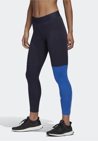 Femme portant des leggings taille haute noir et bleu et des chaussures de course noires, se tenant avec les mains relaxées sur les côtés.
