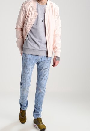 Helle rosa Bomberjacke, grauer Grafik-Sweatshirt, ausgewaschene blaue Jeans mit schwarzen Kritzelmustern und olivgrüne Sneaker.